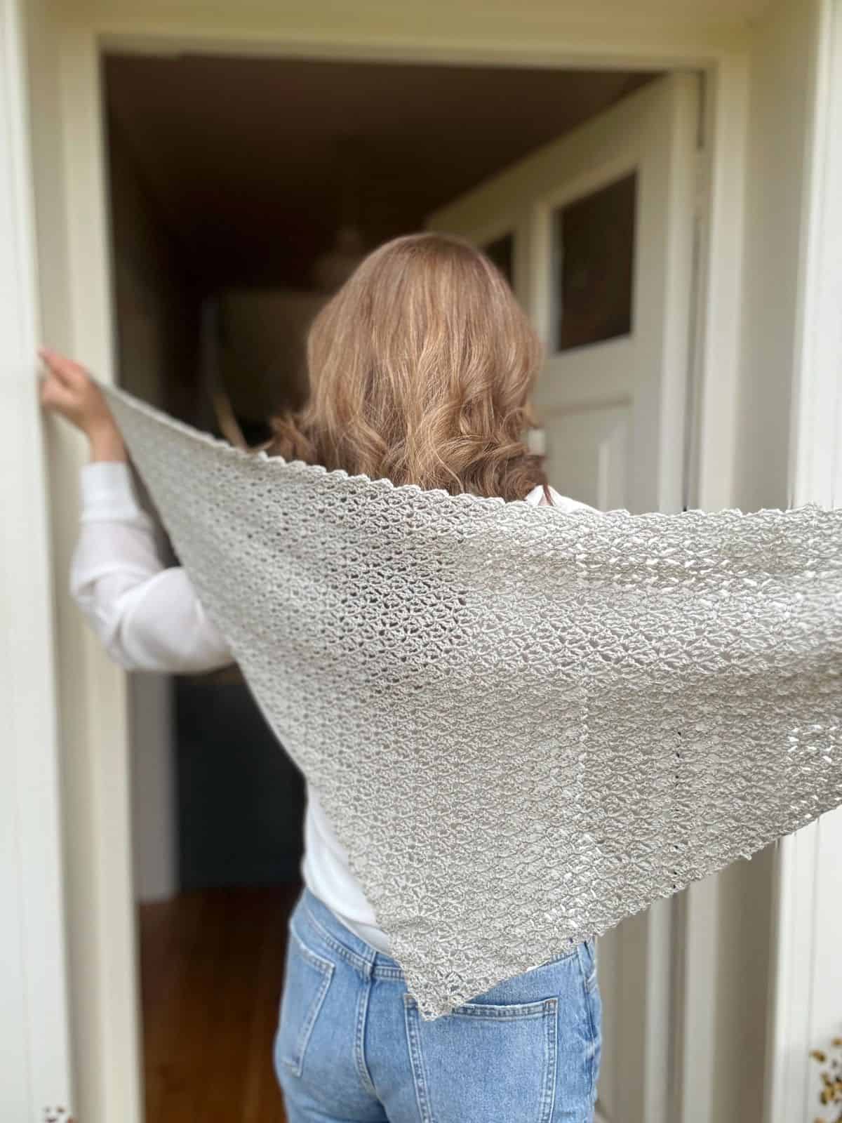 A person with wavy hair, wearing jeans and a white top, stands in a doorway holding out a light gray shawl made using a delicate lace crochet shawl pattern.