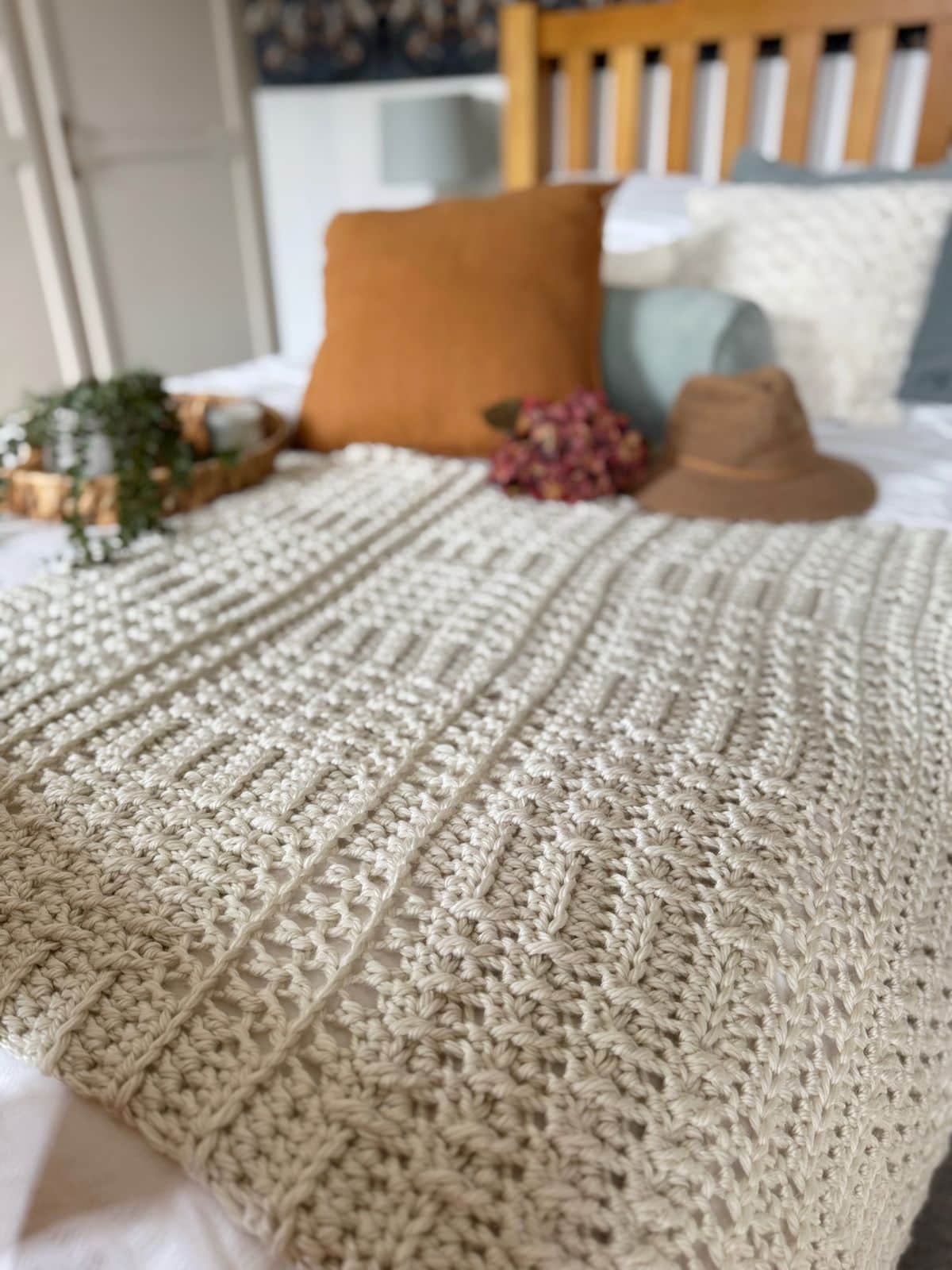 A thick crochet blanket pattern adds texture to the bed, spread alongside pillows, a hat, a tray with a plant, and dried flowers in this softly lit bedroom.
