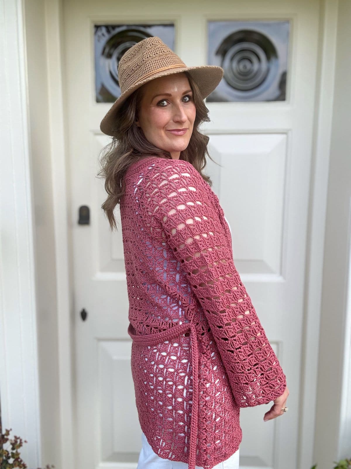 A woman wearing a wide-brimmed hat and a pink summer cardigan crochet pattern stands in front of a white door, looking over her shoulder.