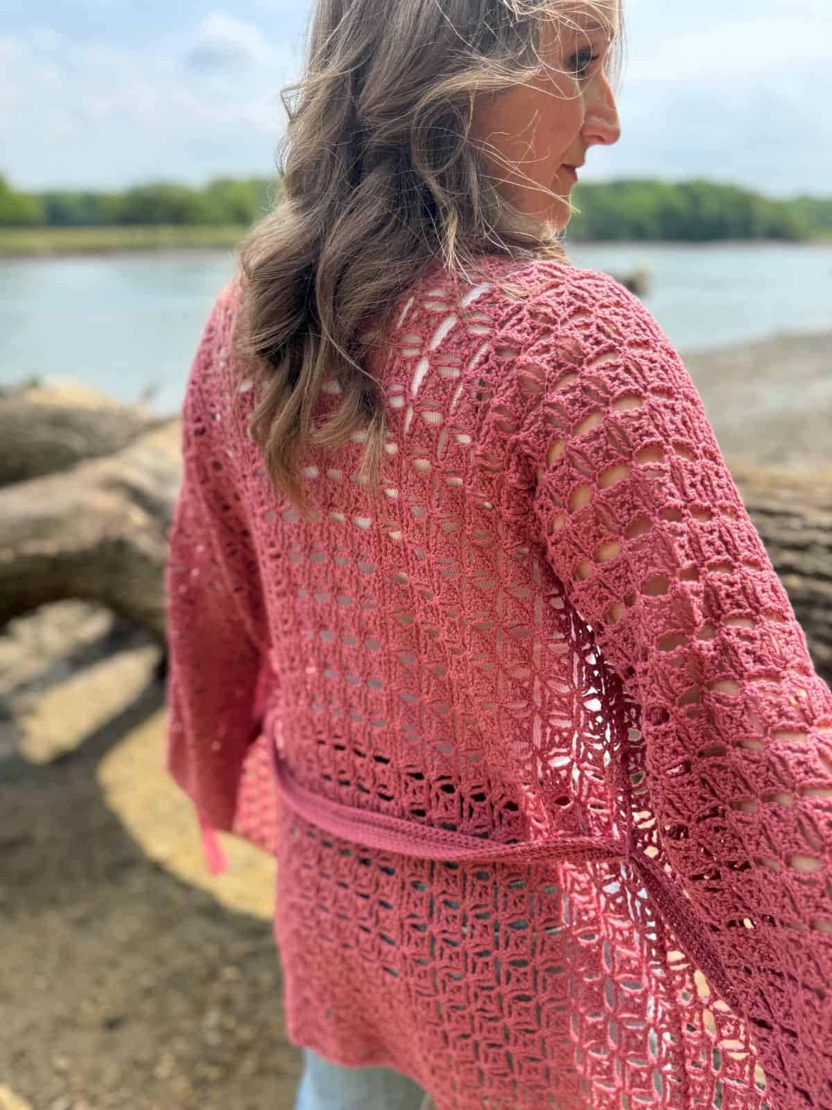 A woman with wavy hair stands outdoors near water, wearing a pink, open-weave summer cardigan crochet pattern with a belt.