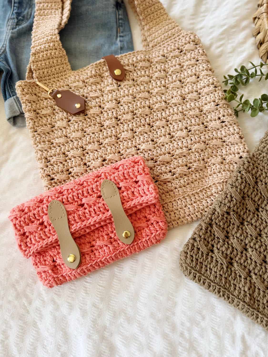 A beige crocheted tote bag with leather accents and a pink crocheted clutch with leather straps lie on a textured white surface, beside a brown piece perfect for trying your next crochet cross body bag free pattern.