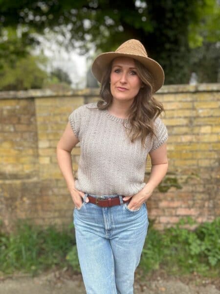 A woman wearing a beige crochet summer top pattern, blue jeans, and a wide-brimmed hat stands outdoors in front of a yellow brick wall with greenery in the background.