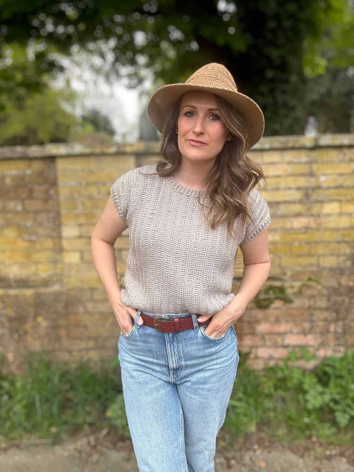 A woman wearing a beige crochet summer top pattern, blue jeans, and a wide-brimmed hat stands outdoors in front of a yellow brick wall with greenery in the background.