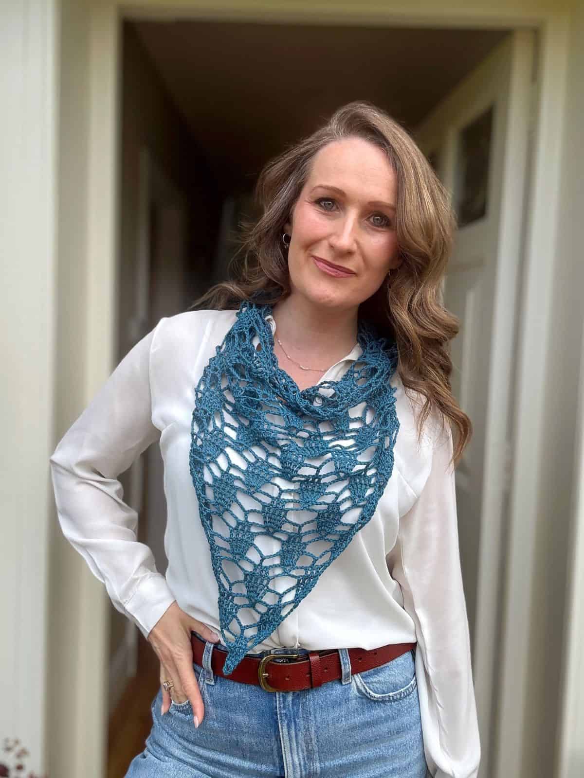 Woman standing indoors wearing a white blouse, blue jeans, brown belt, and a blue bandana scarf crochet pattern draped around her neck, with one hand on her hip and a neutral facial expression.