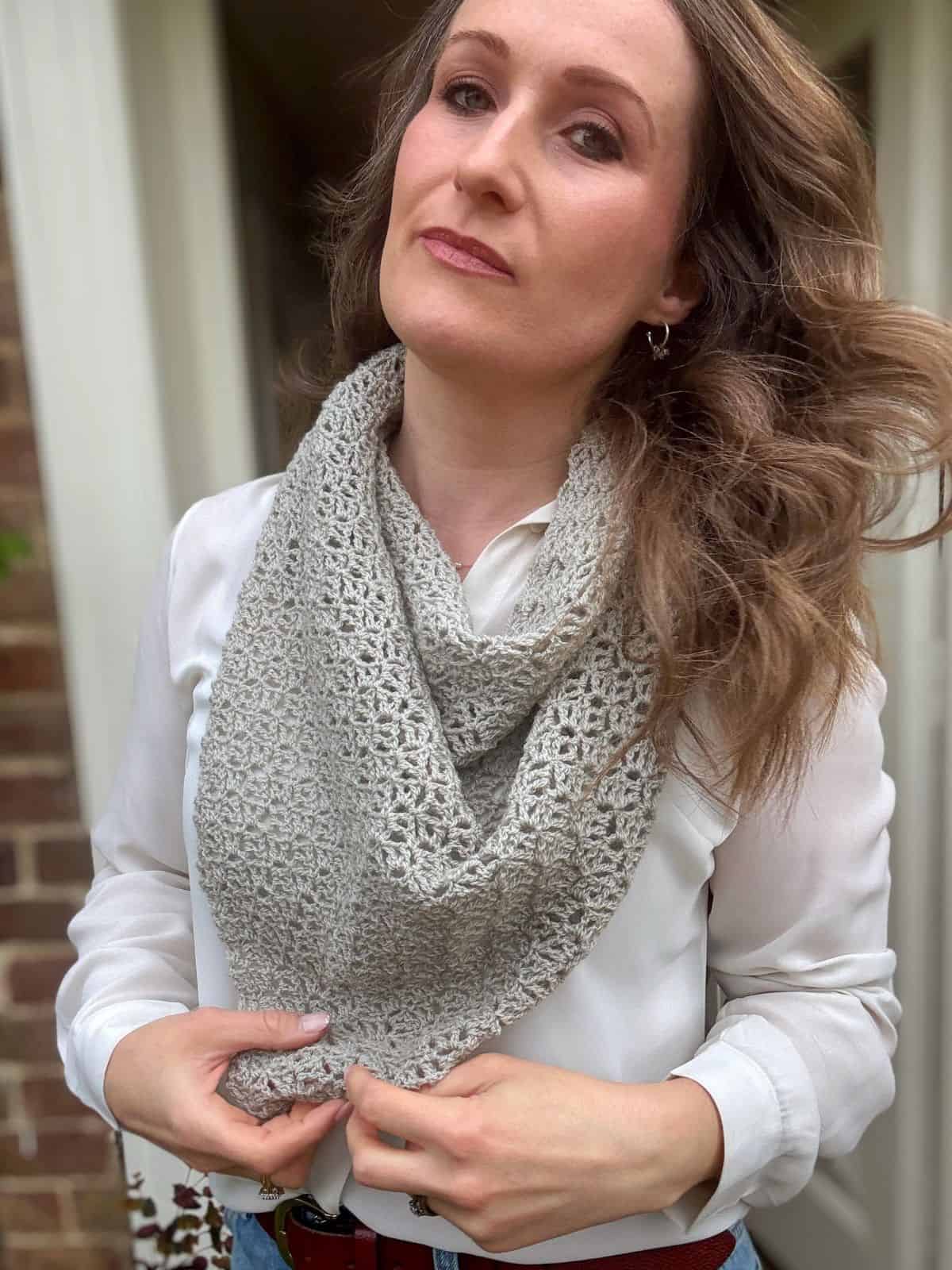 Woman wearing a white blouse and a light gray scarf made from a lace crochet shawl pattern, standing in front of a brick wall with one hand holding the scarf.
