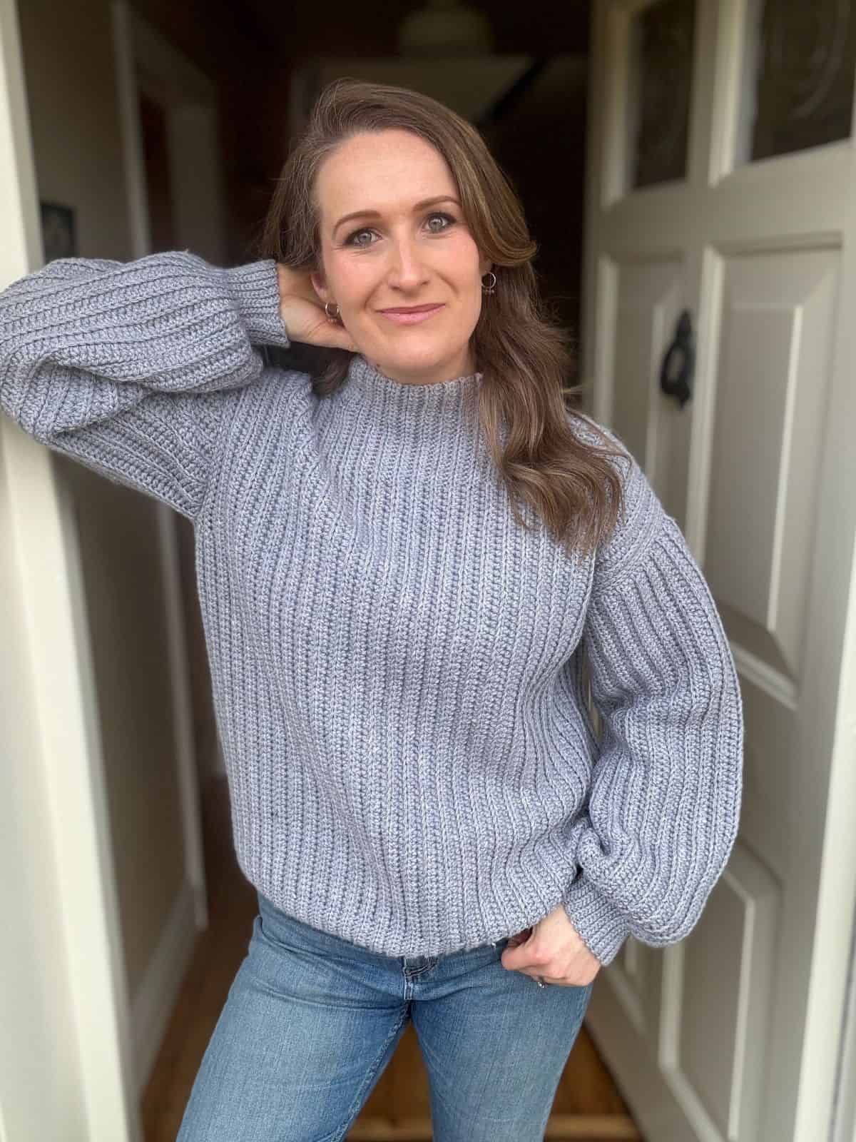 A woman wearing a gray crochet ribbed sweater pattern and blue jeans stands in a doorway with one hand behind her head, looking at the camera.