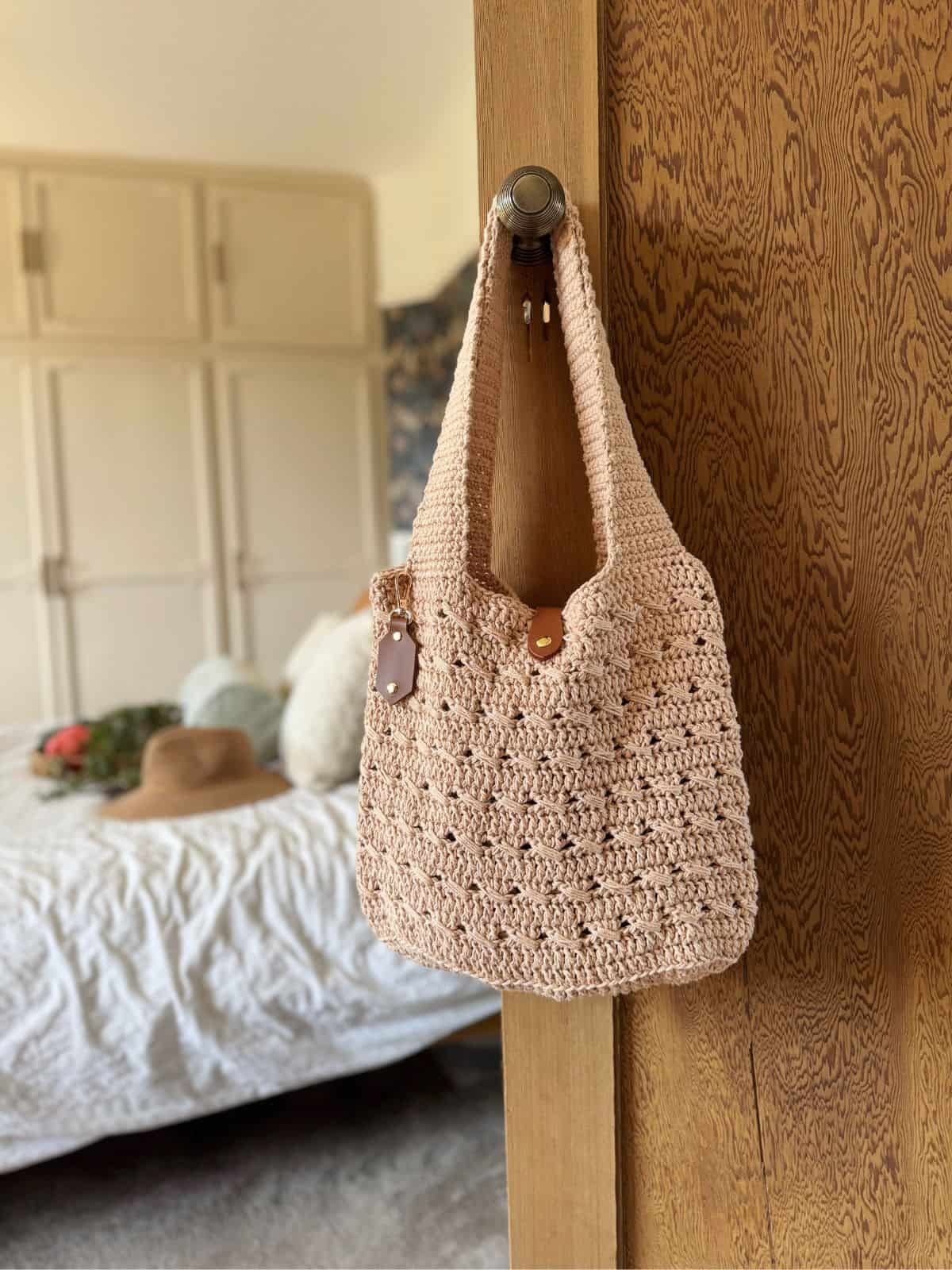 A beige crocheted tote bag made from a free crochet market bag pattern with leather accents hangs on a wooden door handle; a bed, hat, and pillows are visible in the softly lit background.