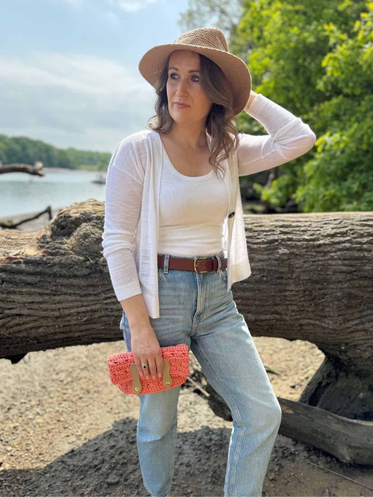 Woman wearing a hat, white cardigan, white top, and jeans stands outdoors by a large fallen tree, holding a pink crochet clutch bag pattern, with trees and water in the background.