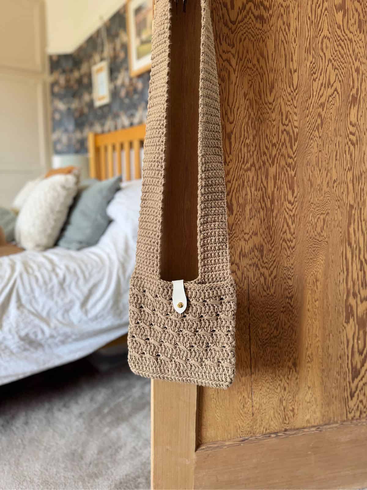 A beige crochet cross body bag, made from a free pattern with a button closure, hangs on a wooden bed frame in a bedroom with a neatly made bed and patterned wallpaper.