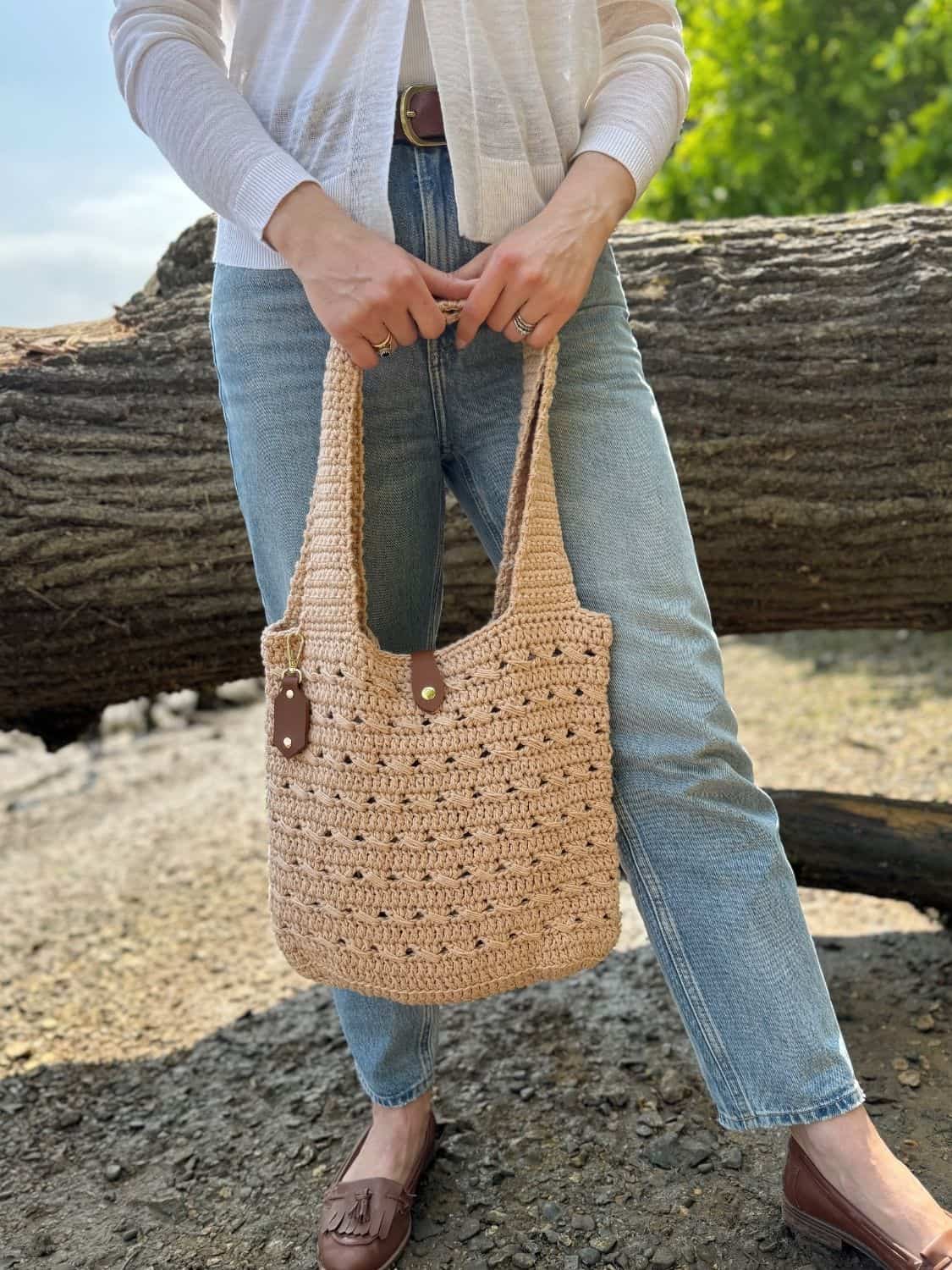 A person in light jeans and a white top holds a beige crochet tote bag with brown leather accents, styled like a crochet cross body bag free pattern, standing in front of a large fallen tree outdoors.