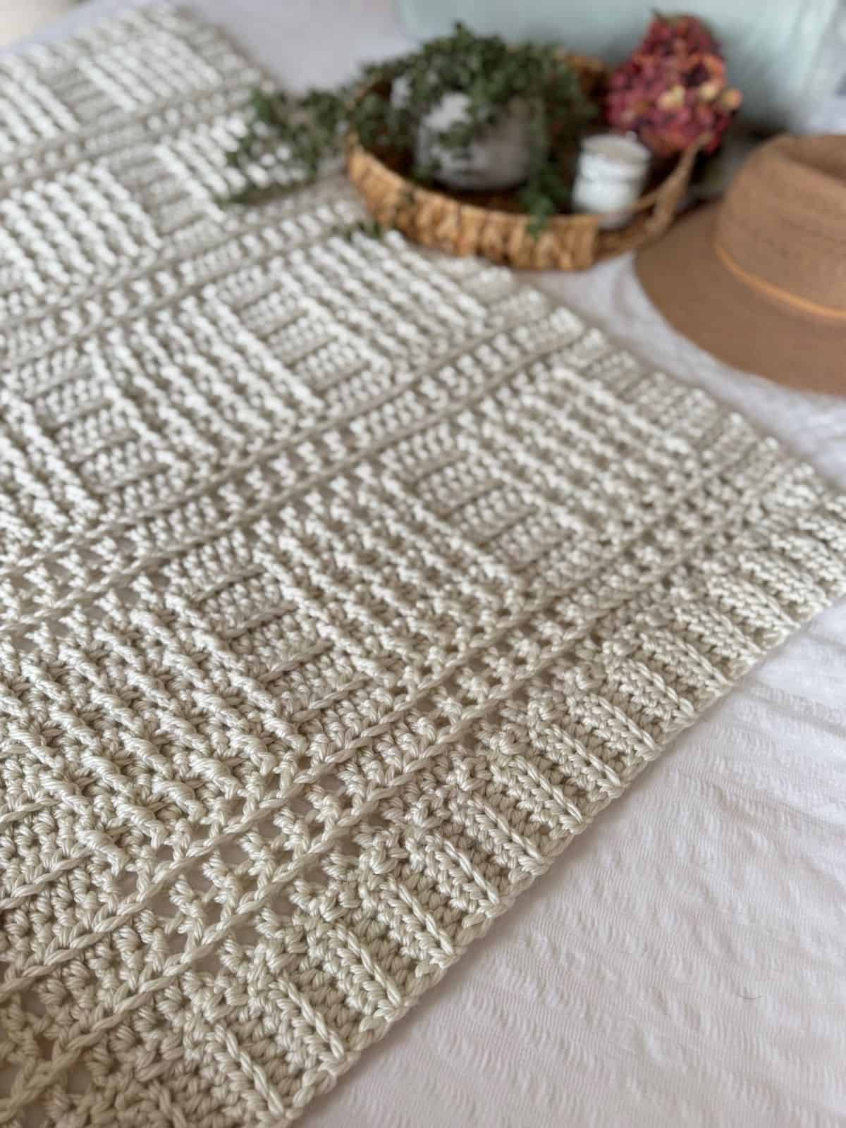 A cream-colored blanket made with a thick crochet blanket pattern features geometric designs and lies on a bed beside a woven tray holding plants, candles, and a brown hat.