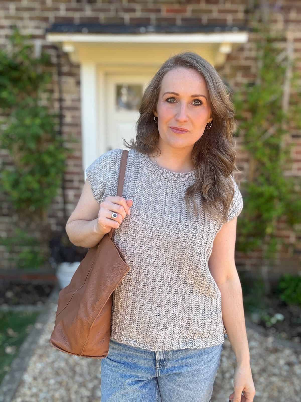 A woman with wavy brown hair stands outside in front of a brick wall, wearing a blue jeans and holding a brown shoulder bag, paired with a stylish crochet summer top pattern.