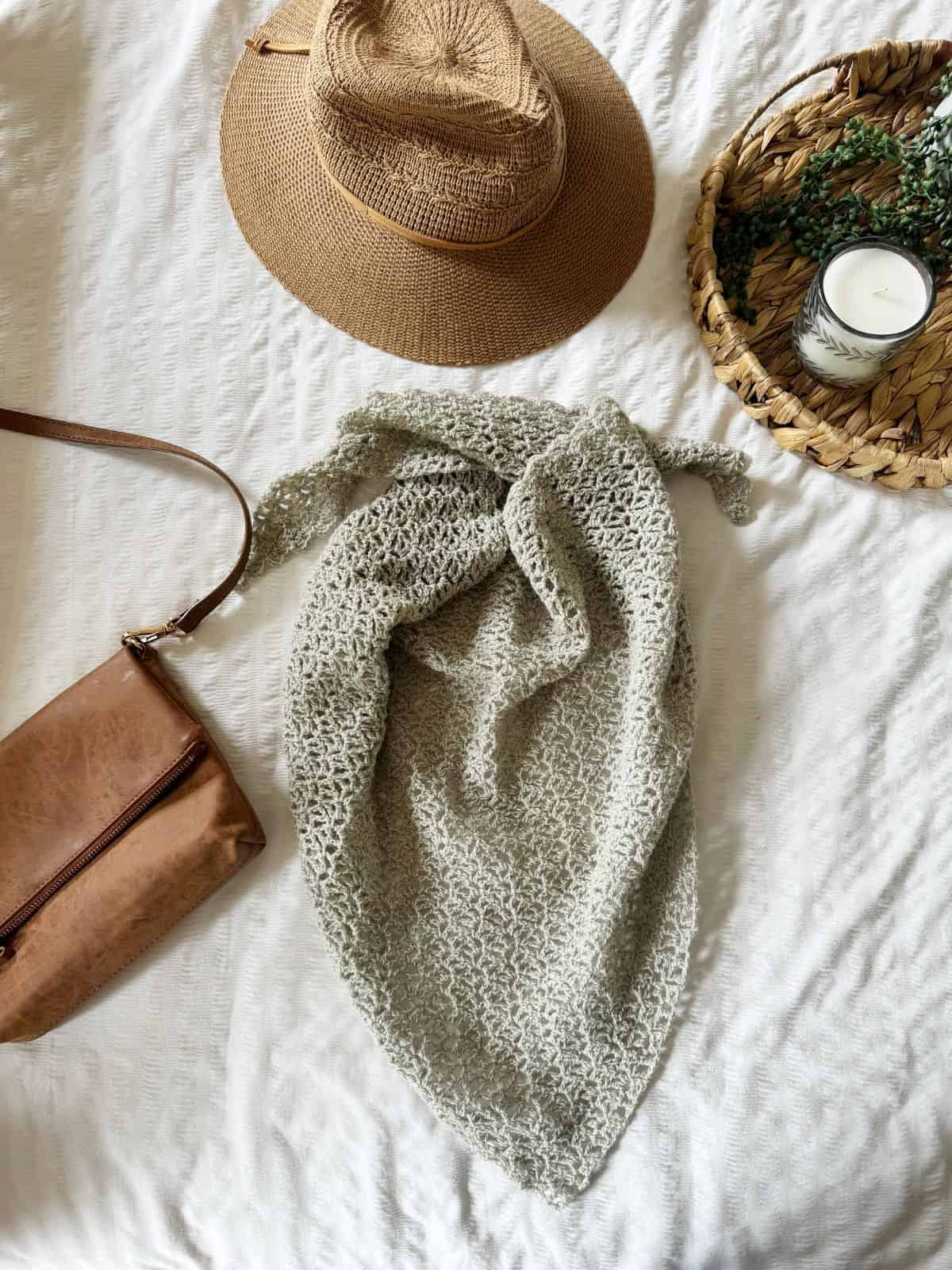 A straw hat, a brown purse, a lace crochet shawl pattern in light gray, and a round tray with a candle and greenery on a white textured surface.