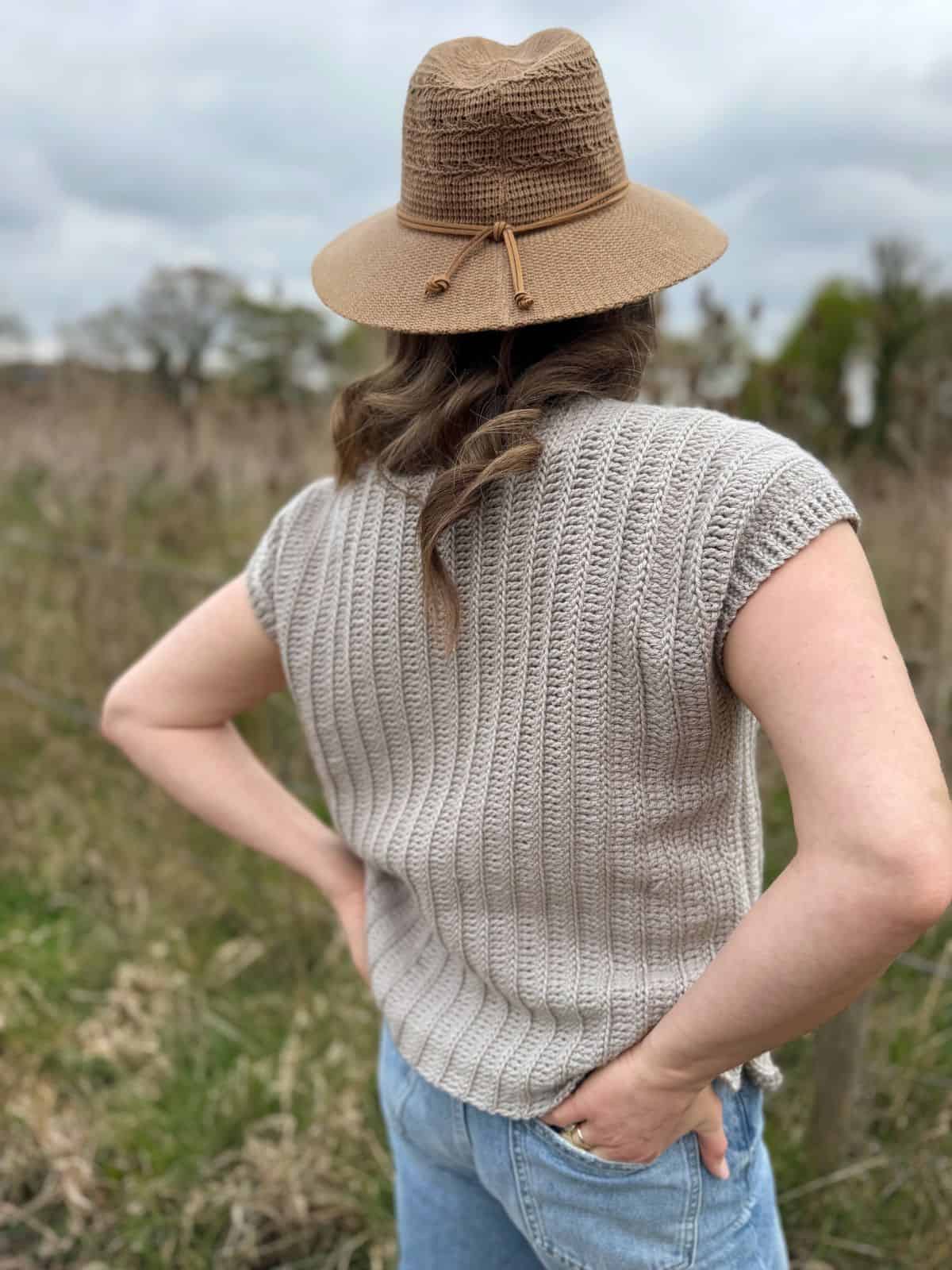 A person wearing a beige crochet summer top pattern, light blue jeans, and a brown hat stands outdoors with hands on hips, facing away from the camera.