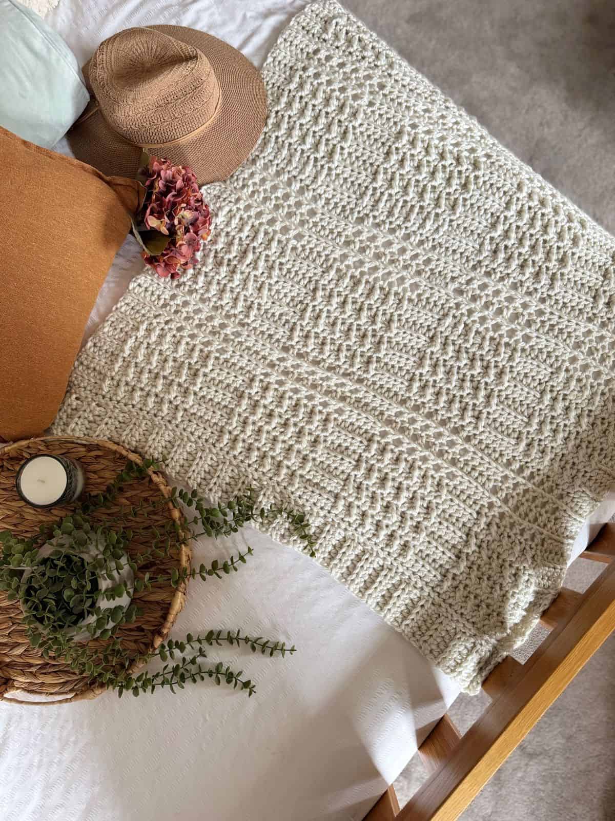 A thick crochet blanket pattern creates a textured cream crochet blanket, spread on a bed next to a brown pillow, straw hat, dried flowers, a candle, and a woven tray with greenery.