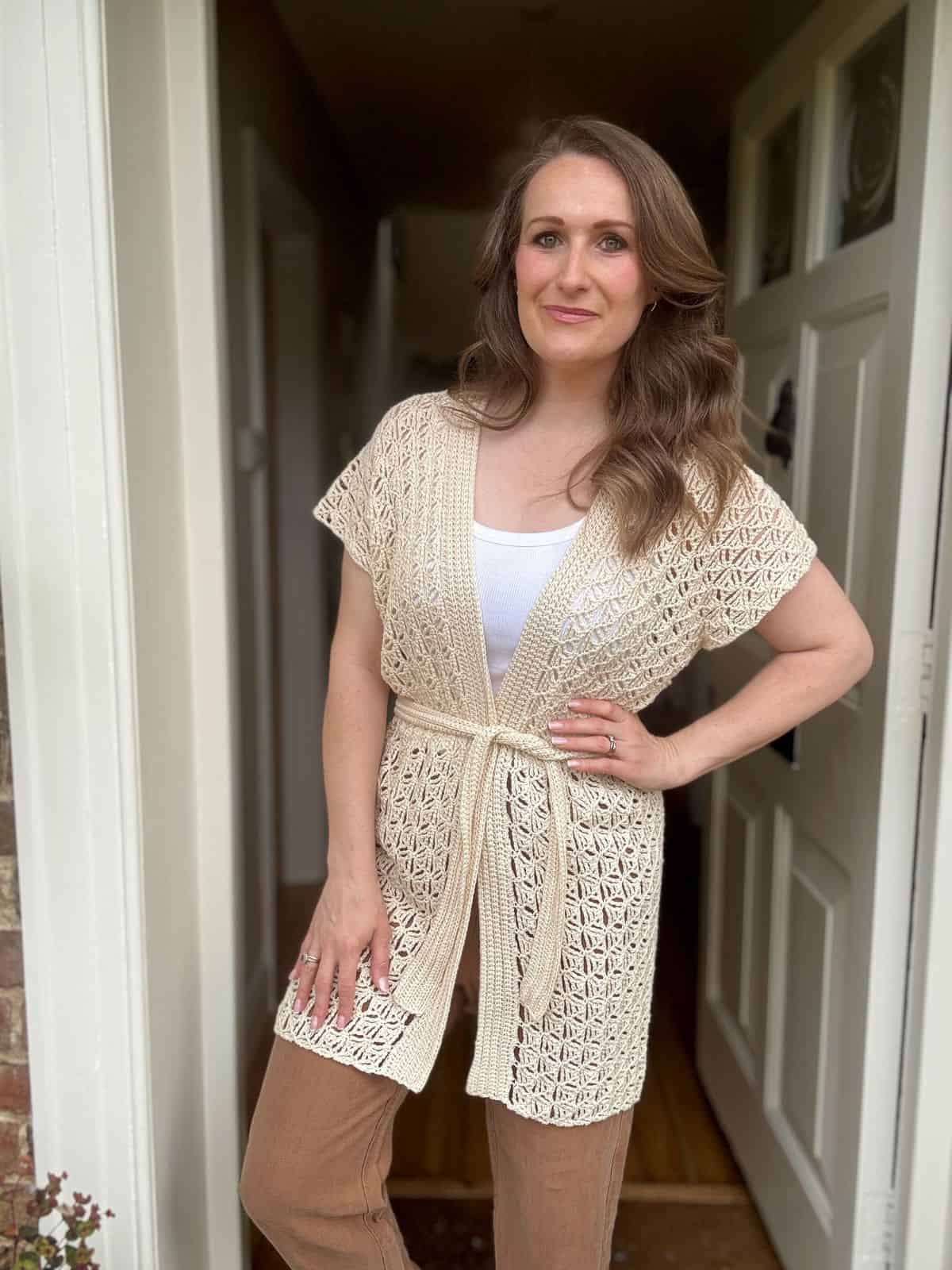 Woman standing in a doorway, wearing a cream-colored summer cardigan crochet pattern with a belt, a white top, and light brown pants, with one hand on her hip.