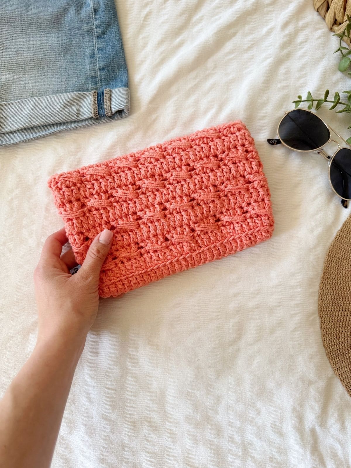 A hand holding a pink crocheted clutch rests on a white textured surface beside denim shorts, sunglasses, and part of a straw hat—perfect inspiration for your next crochet clutch bag pattern.