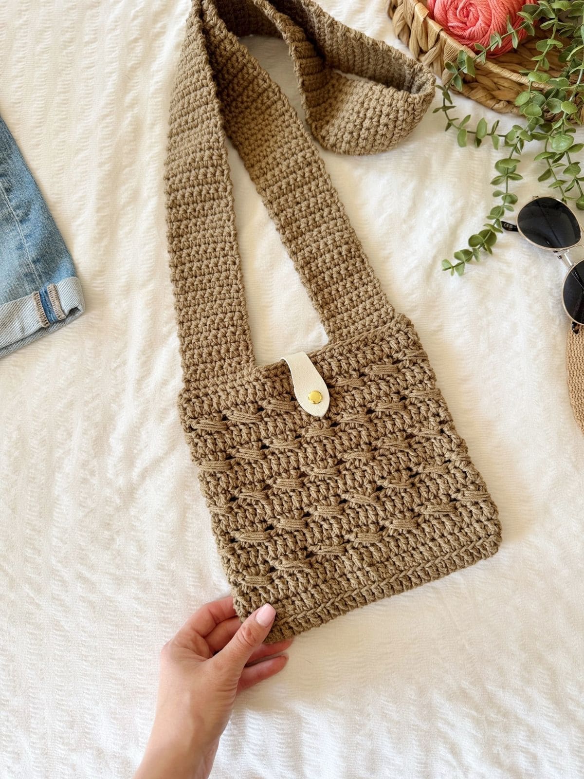 A hand holds a beige crochet cross body bag with a wide strap and white button closure, placed on a white textured surface near sunglasses, greenery, denim fabric, and featuring a crochet cross body bag free pattern.