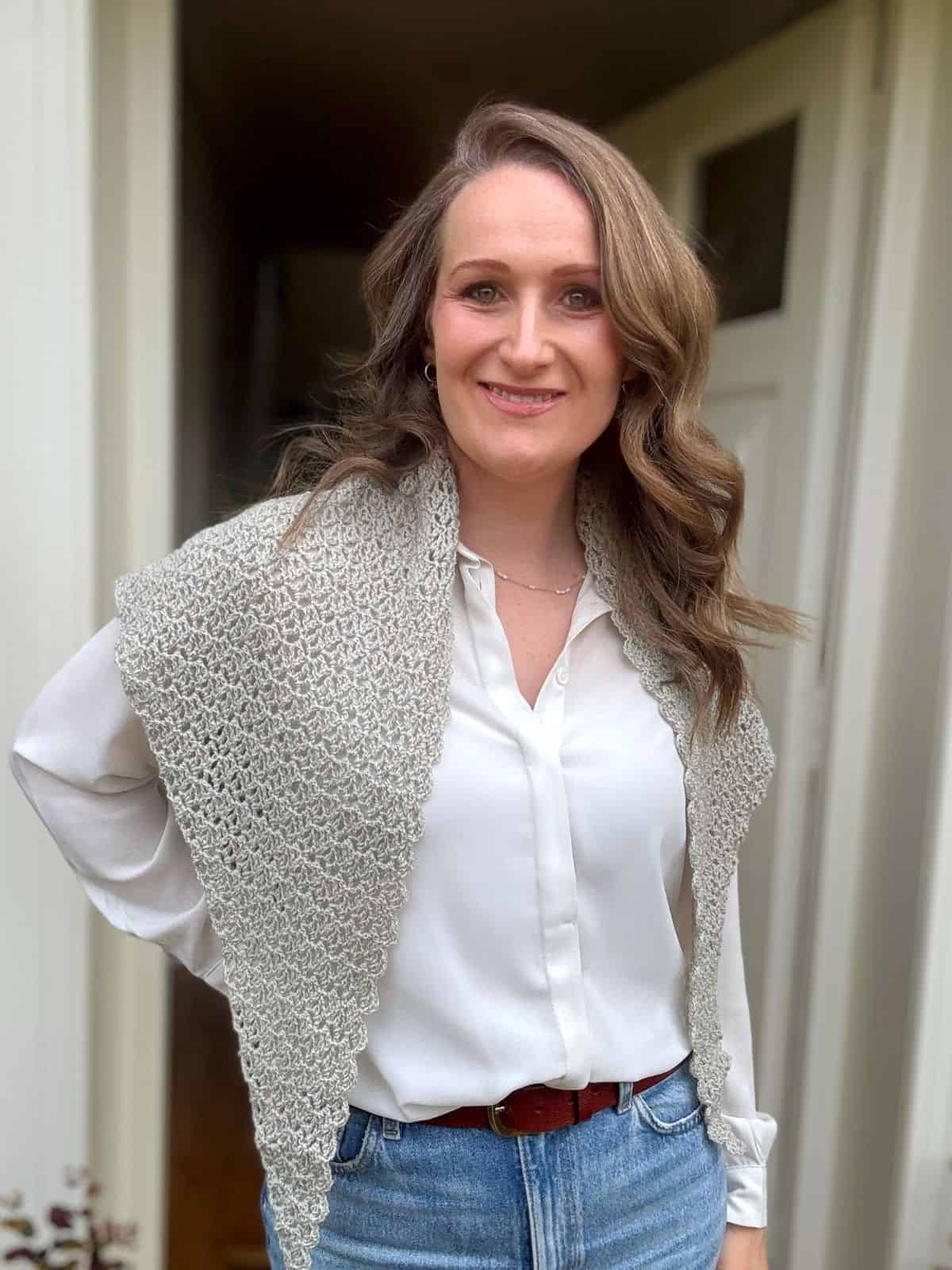 A woman with wavy hair stands indoors wearing a white blouse, blue jeans, and a delicate lace crochet shawl pattern draped over her shoulders.