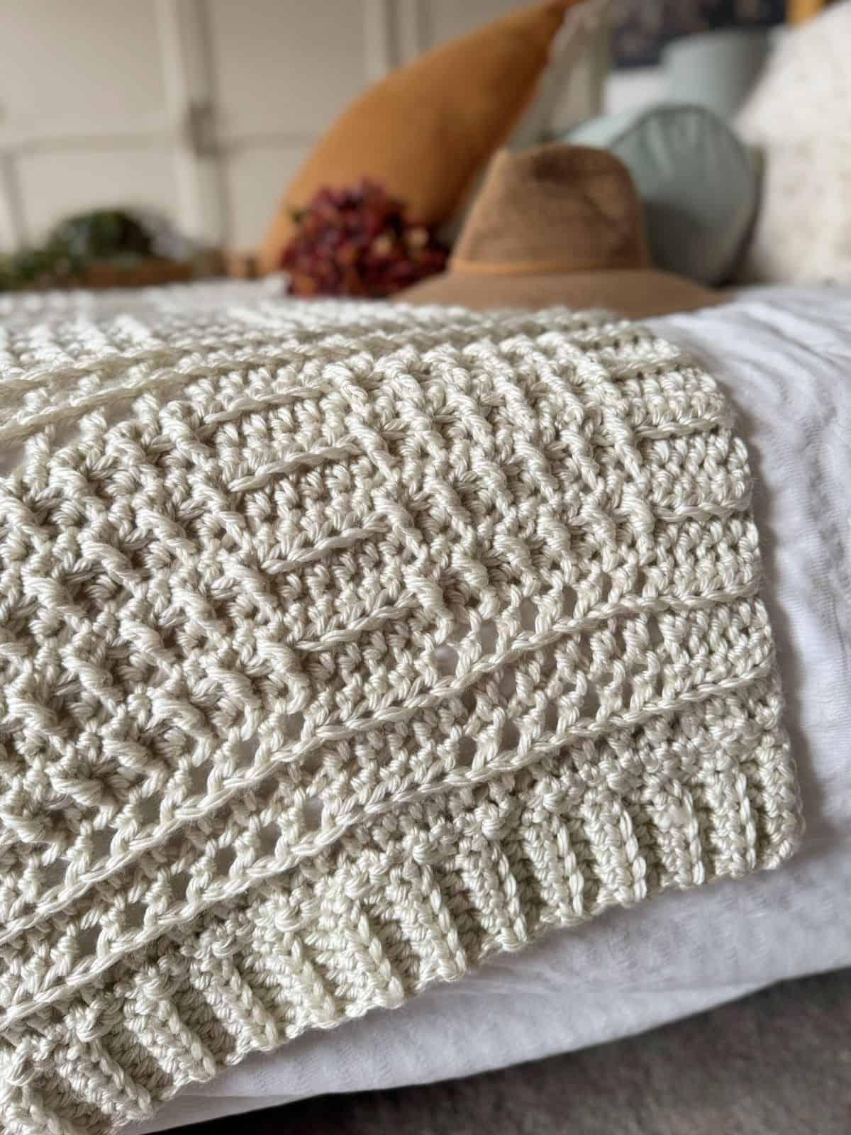 A close-up of a cream-colored thick crochet blanket pattern draped over the edge of a bed, with pillows and a brown hat in the background.
