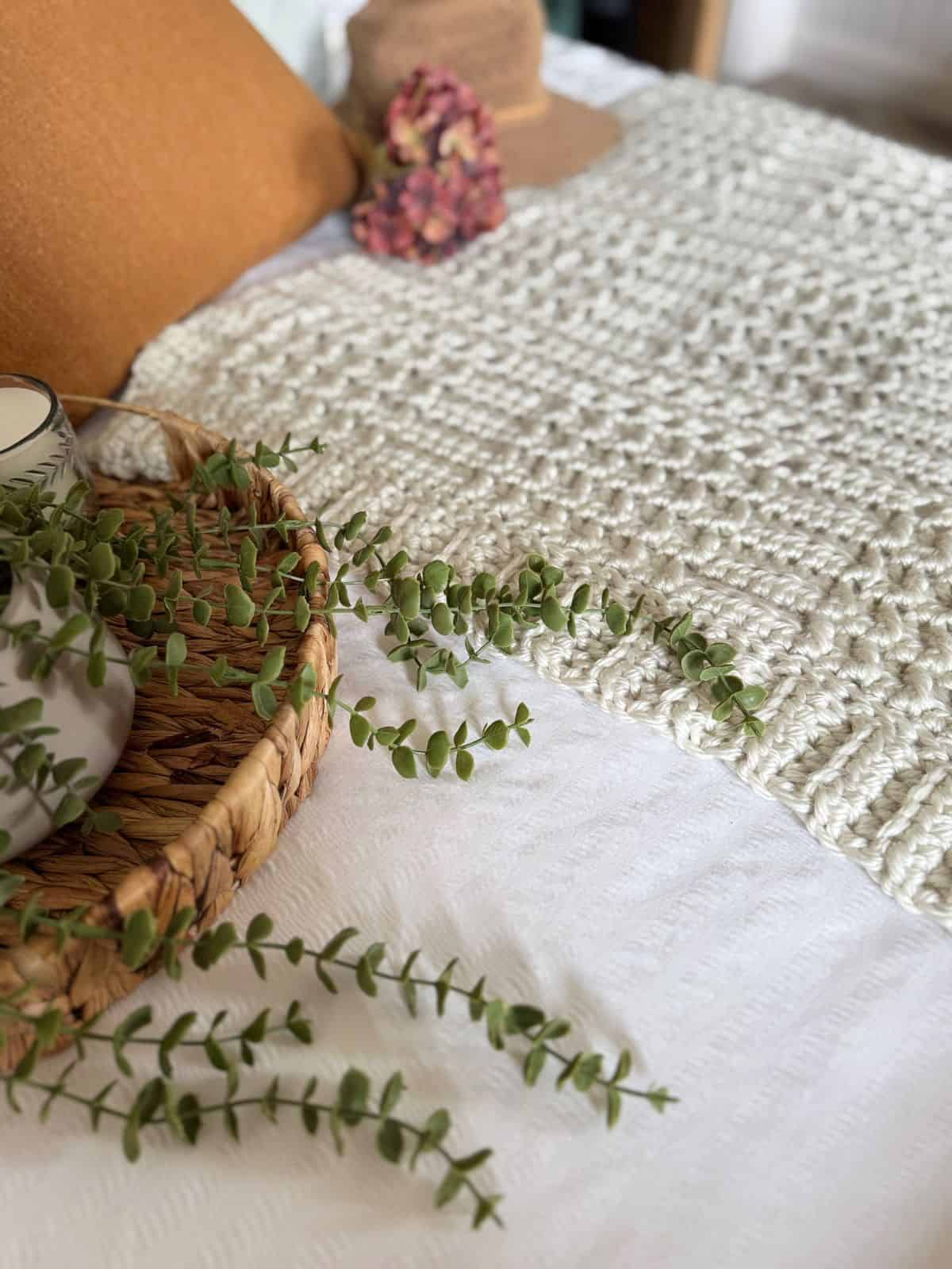 A woven tray with green foliage rests atop a thick crochet blanket pattern in cream, set on a bed with orange pillows and a hat adorned with dried flowers in the background.