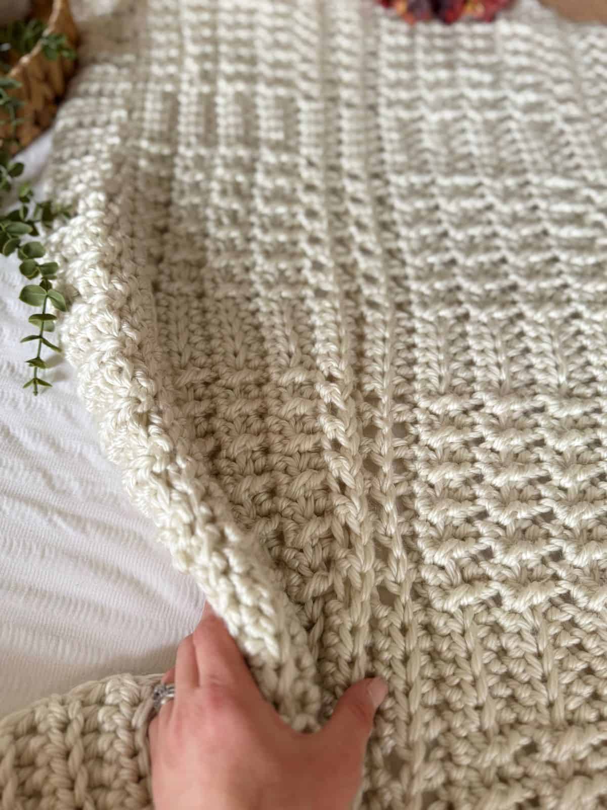 A hand holds up the edge of a chunky, cream-colored blanket, made with a thick crochet blanket pattern, laid out on a white surface with green foliage nearby.
