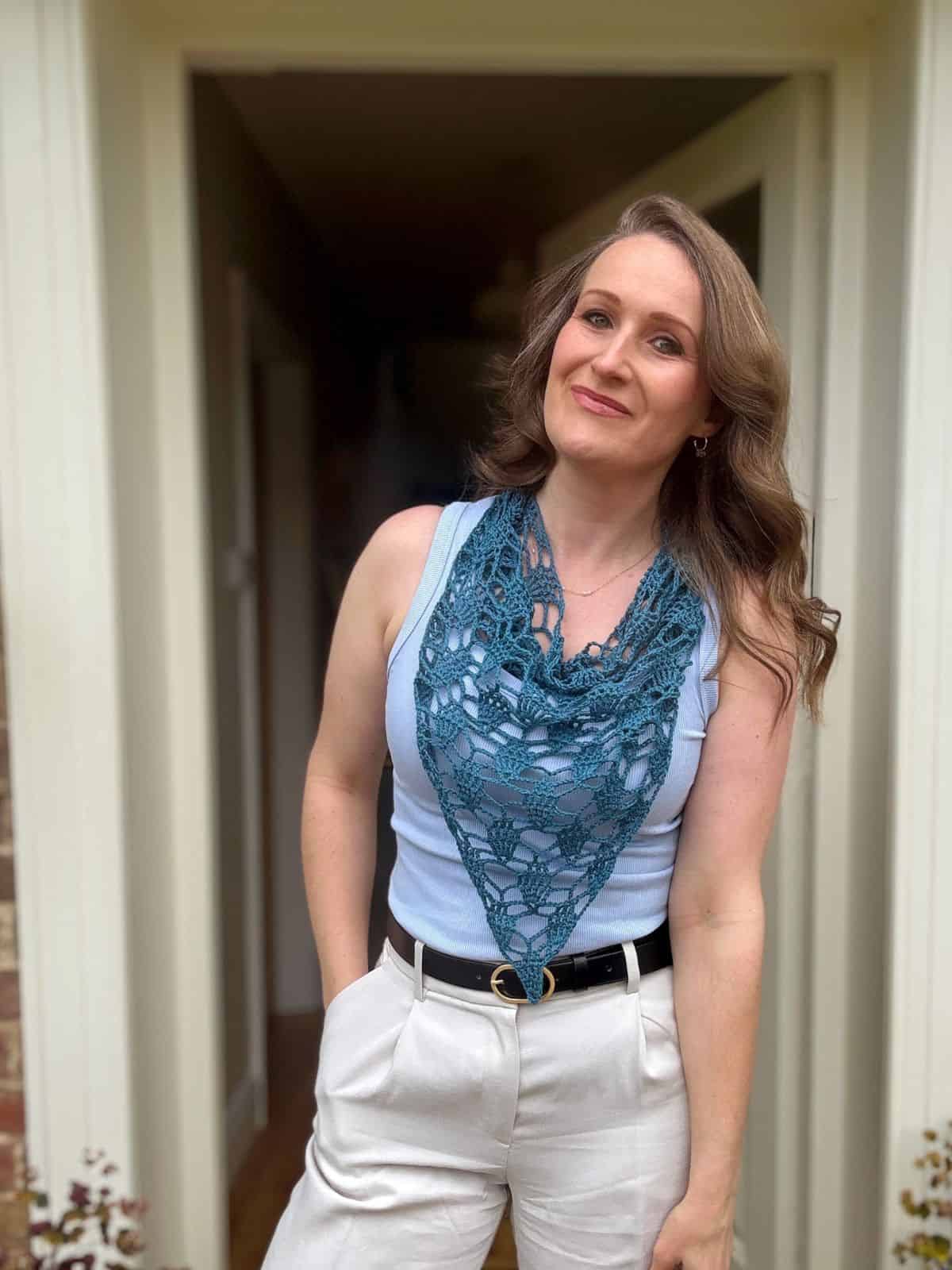 Woman standing in a doorway, wearing a light blue sleeveless top, white pants, black belt, and a teal bandana scarf crochet pattern draped around her neck, with one hand in her pocket.