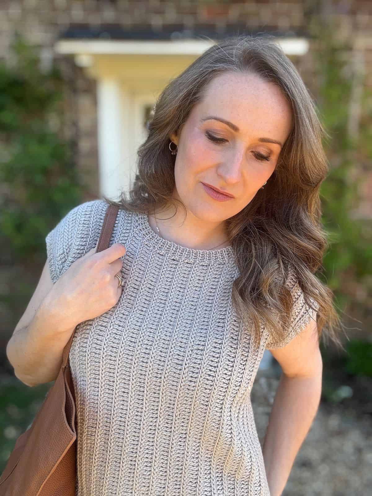 A woman with wavy light brown hair wearing a short-sleeved crochet summer top stands outside in front of a brick wall, looking down with a neutral expression.