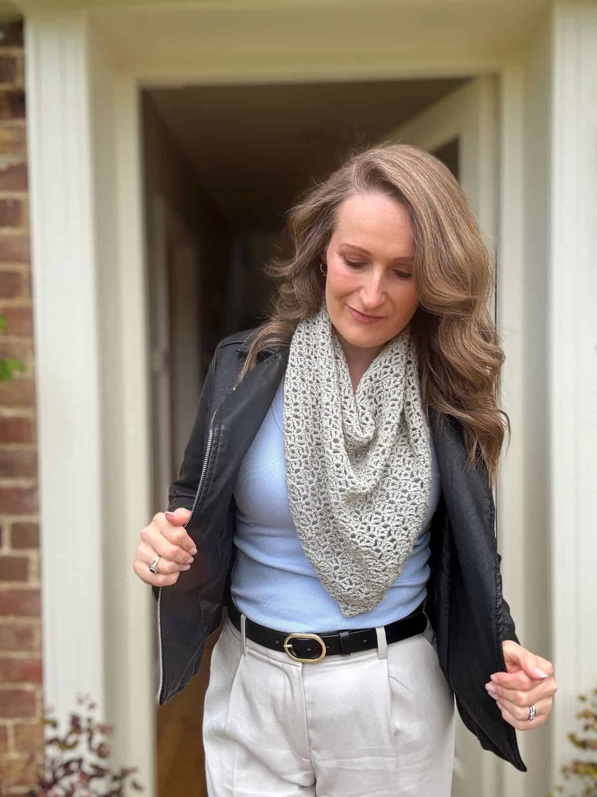 Woman standing in a doorway, wearing a black jacket, light blue top, white pants, and a lace crochet shawl pattern scarf, looking down and adjusting her jacket.