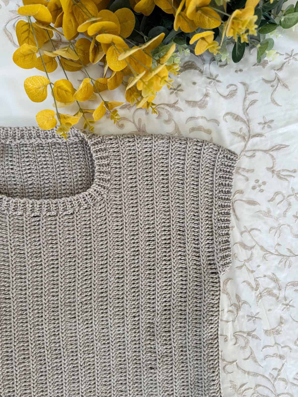 A close-up of a beige ribbed crochet top on embroidered white fabric, with yellow and green artificial flowers in the upper left corner, evokes the delicate texture of a crochet summer top pattern.