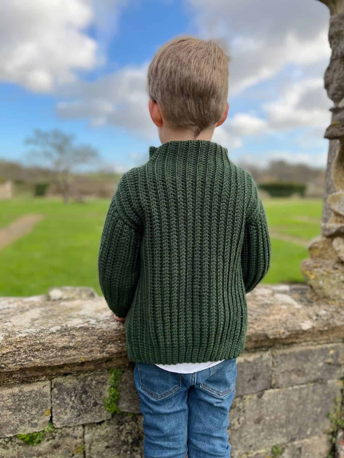 A young child with short blonde hair, wearing a green crochet sweater—perfect inspiration for easy crochet sweater patterns for beginners—and blue jeans, stands outdoors facing away, looking over a stone wall at a grassy area under a partly cloudy sky.