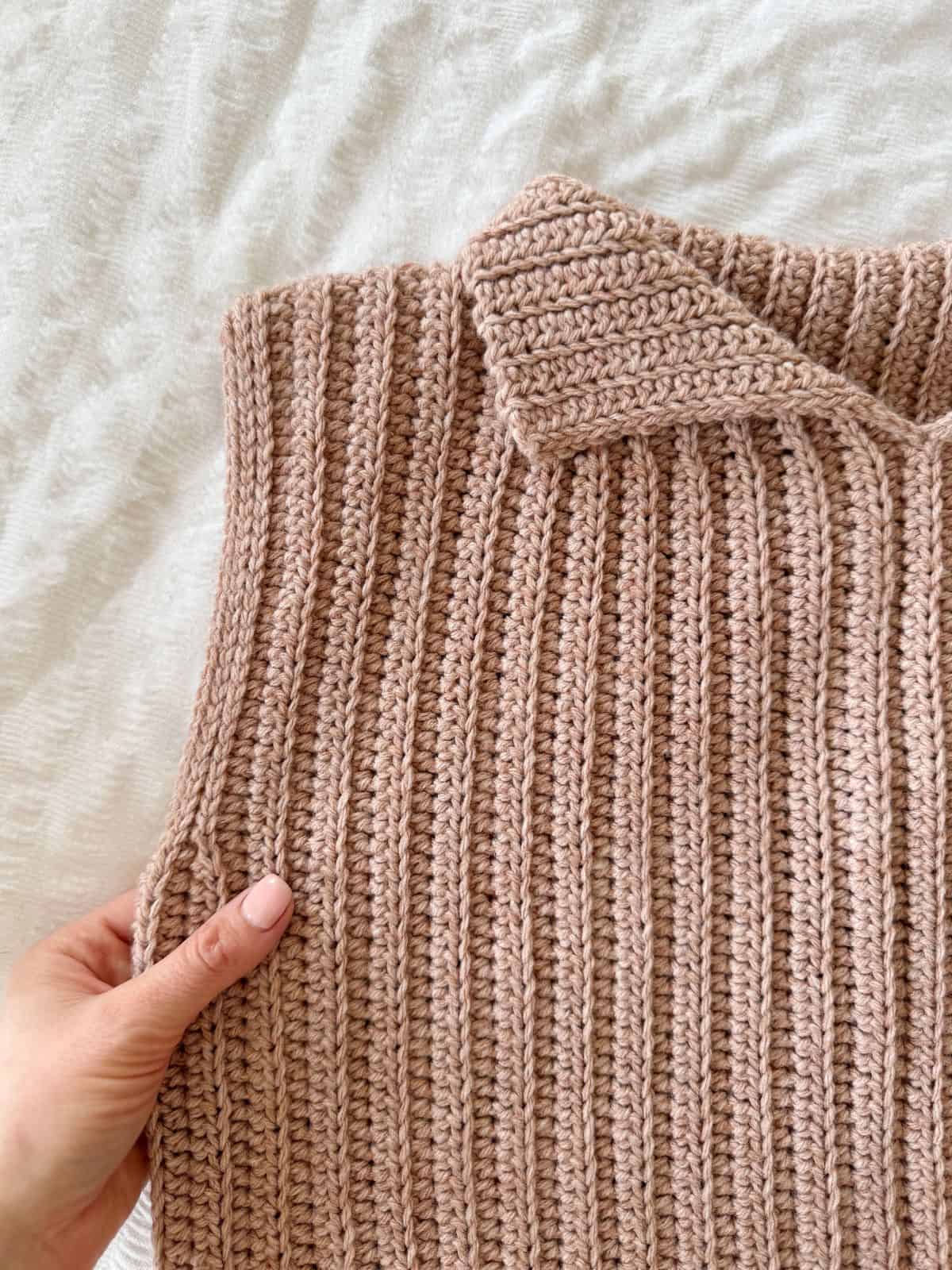 A hand holds a beige, ribbed, crocheted garment with a collar—resembling a modern crochet vest pattern—displayed on a textured white surface.