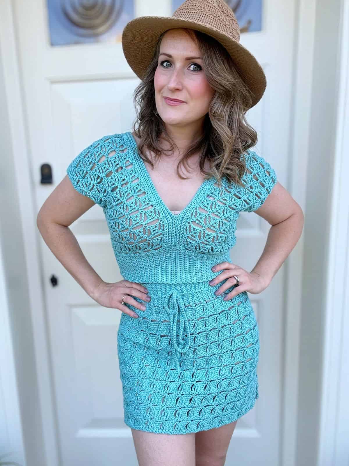Woman wearing a light blue summer cardigan crochet pattern dress and a tan hat stands in front of a white door with her hands on her hips.
