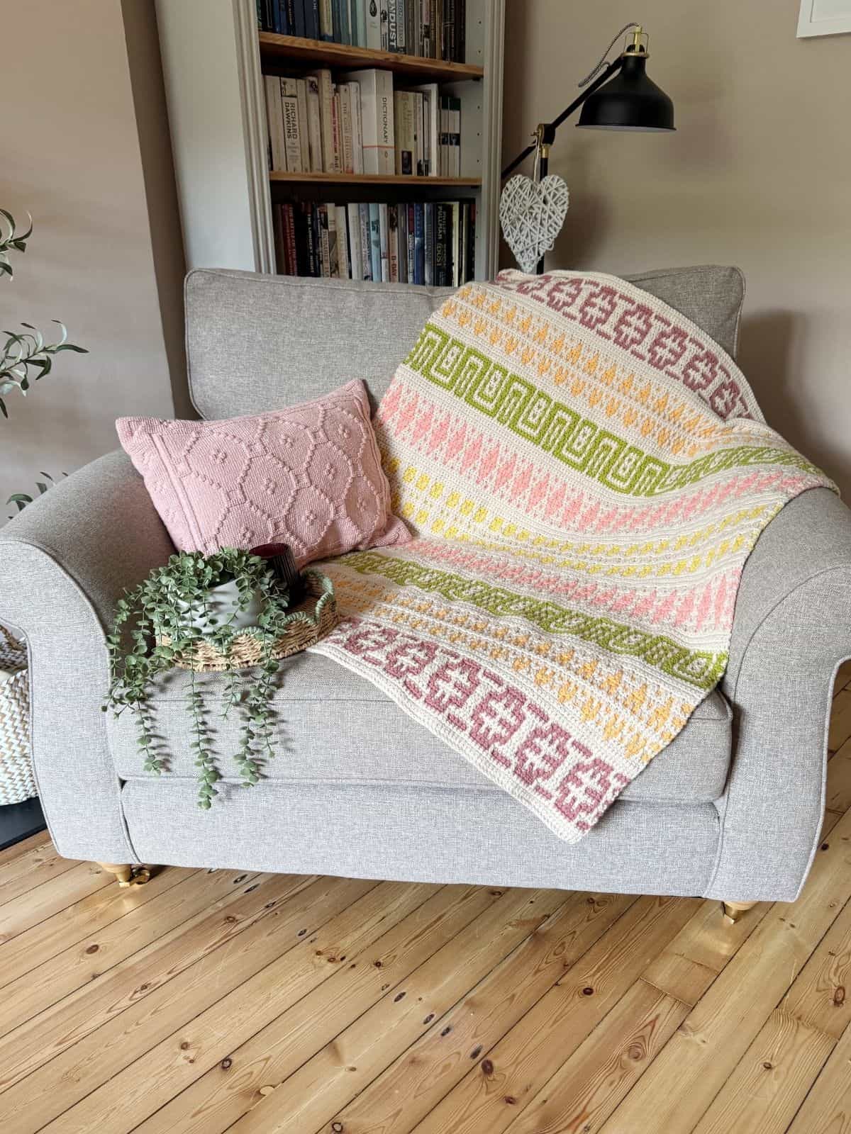 A beige armchair with a pink pillow, a vibrant mosaic crochet blanket pattern, and a potted trailing plant sits on a wooden floor in front of a bookcase.