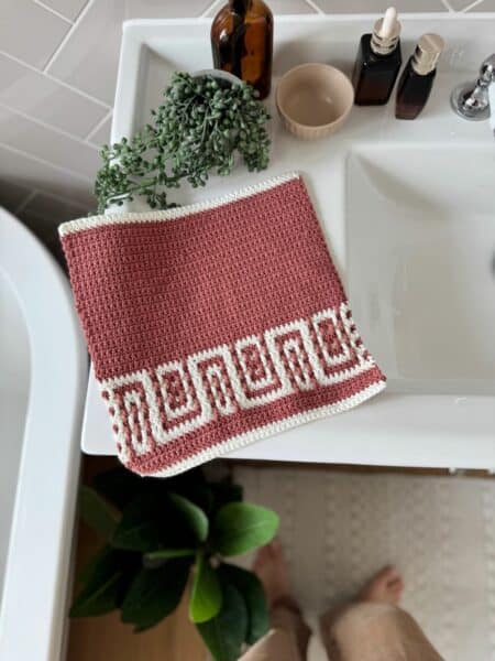 A red and white crochet towel with a geometric border, reminiscent of a free mosaic crochet pattern, rests on a white bathroom sink beside brown bottles, a small dish, and a green plant.