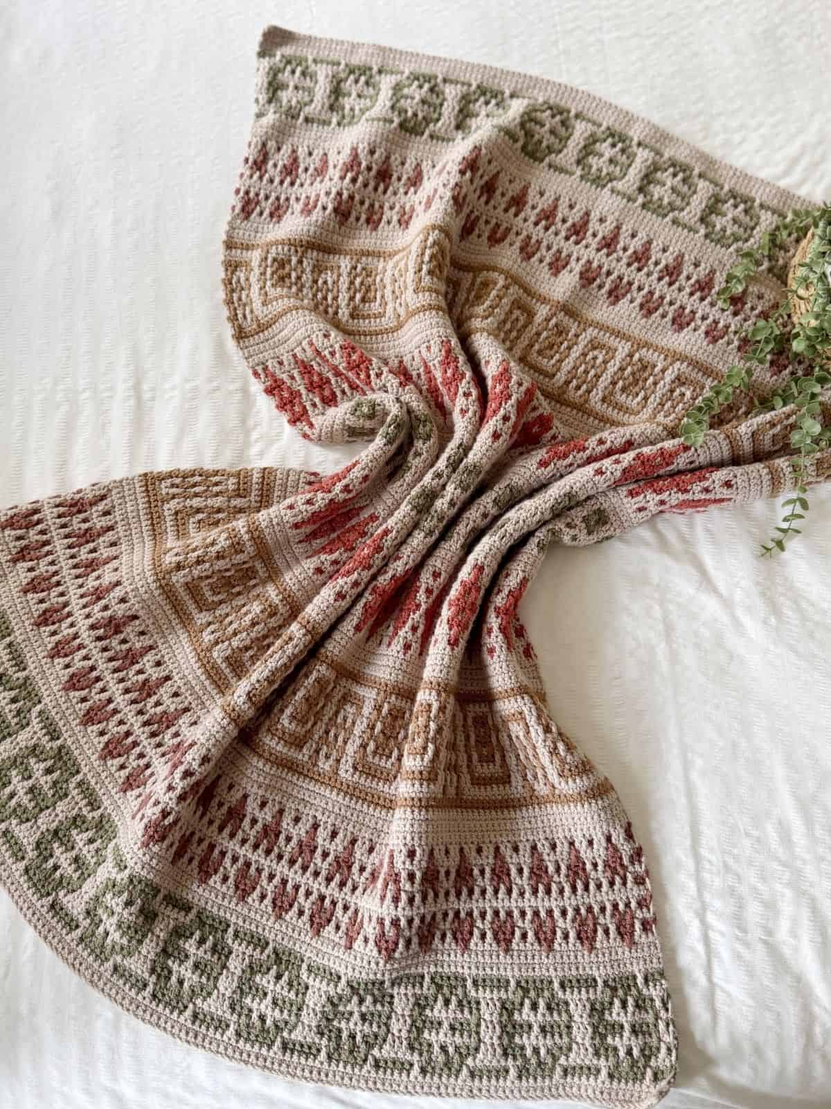 A mosaic crochet blanket pattern with green, brown, and red geometric designs is spread out on a white surface, accented by green plant leaves at the edge.