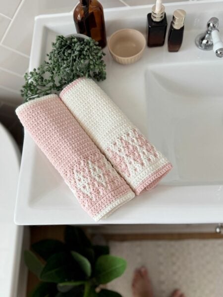 Two folded pink and white towels, crafted with a simple mosaic crochet pattern, rest on a white bathroom sink beside a brown bottle, small bowl, two pump bottles, and a potted plant.