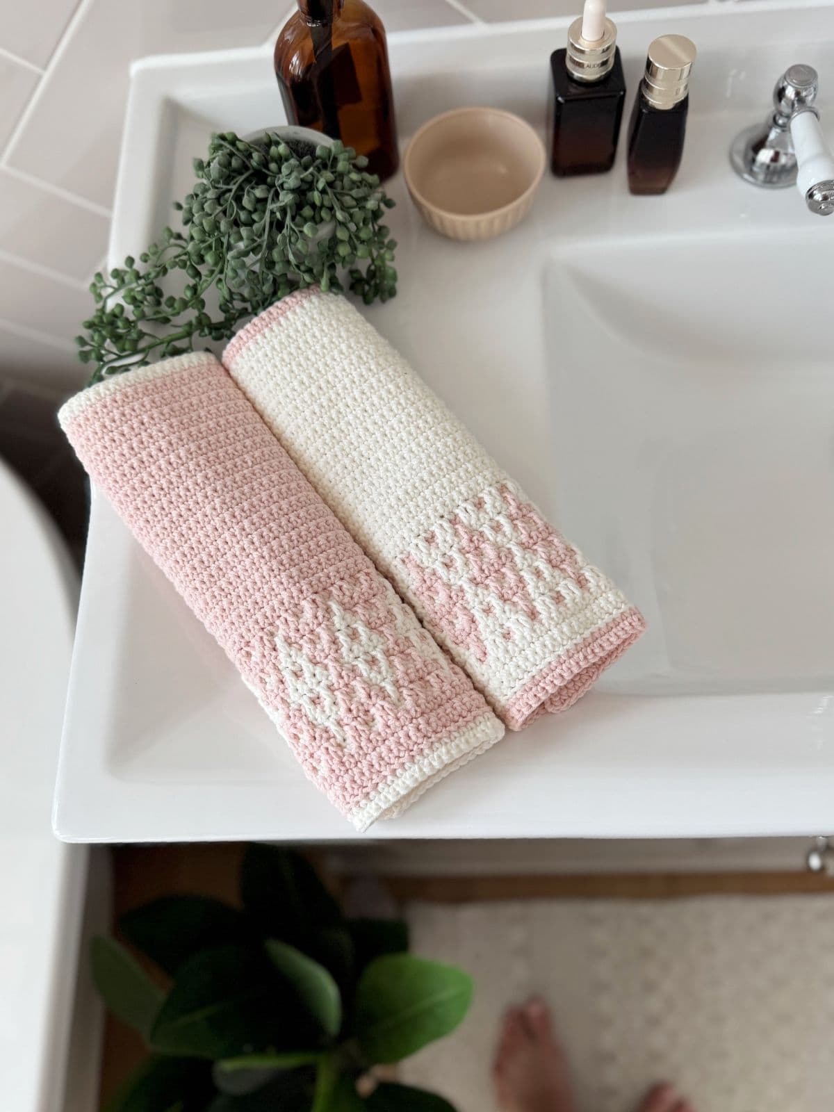 Two folded pink and white towels, crafted with a simple mosaic crochet pattern, rest on a white bathroom sink beside a brown bottle, small bowl, two pump bottles, and a potted plant.
