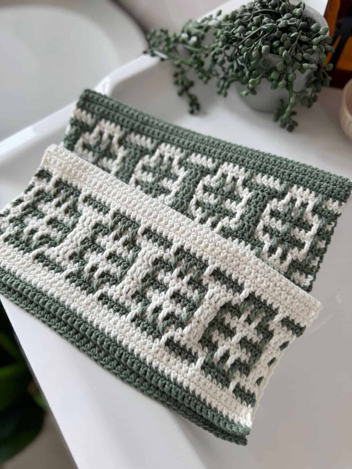 Two rectangular pieces of crochet fabric with geometric patterns in green and white yarn, made using an easy mosaic crochet pattern, displayed on a white surface near a green plant.