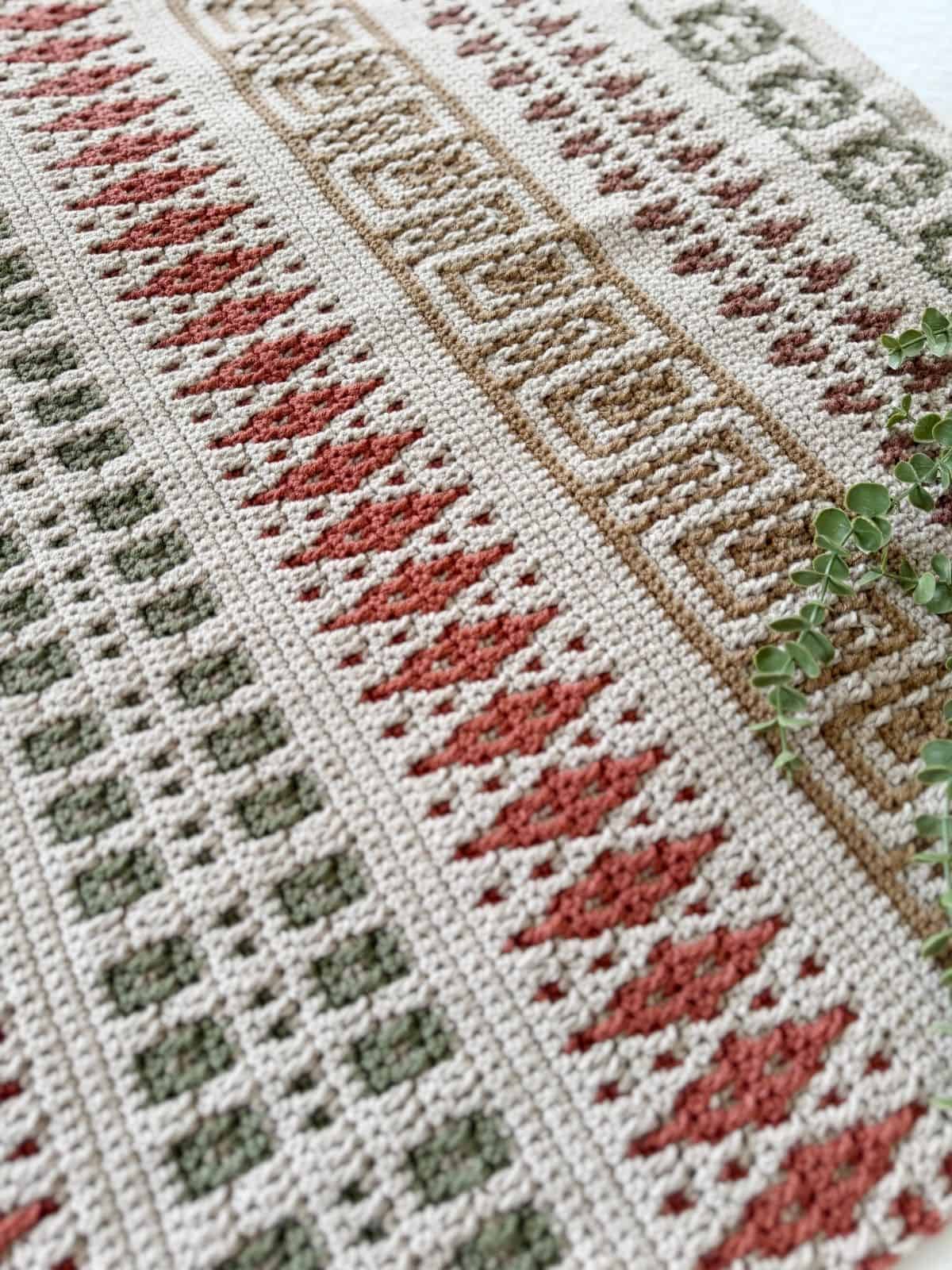 Crocheted fabric with geometric patterns in green, red, and beige tones, displayed flat with a sprig of greenery on the right side—a beautiful example of a mosaic crochet blanket pattern.