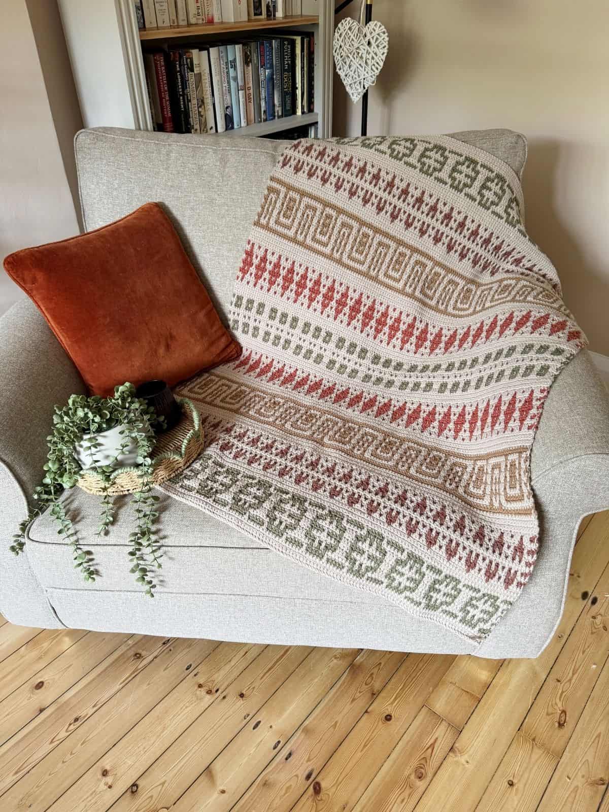 A mosaic crochet blanket pattern is draped over a light gray armchair with a rust-colored pillow and a small potted plant on the armrest; a bookshelf sits in the background.
