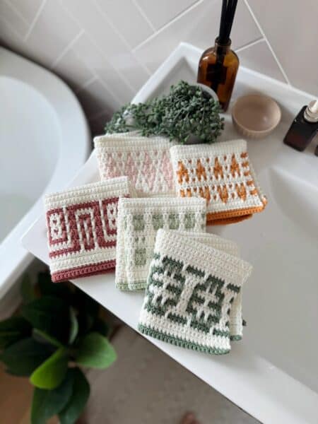 Four crocheted washcloths made with an easy mosaic crochet pattern and featuring geometric designs in different colors are displayed on a white bathroom sink, with plants and toiletries nearby.
