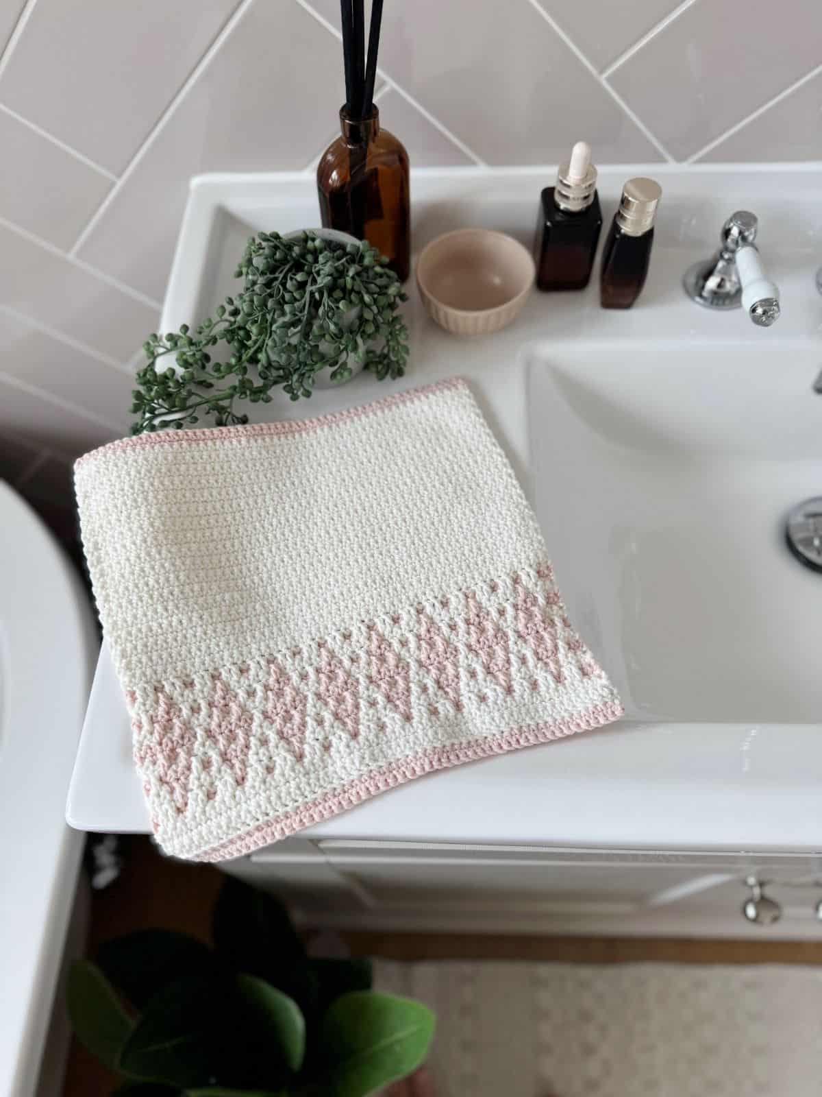 A white and pink crochet hand towel featuring a diamond design, reminiscent of a simple mosaic crochet pattern, is draped over a bathroom sink next to bottles, a bowl, and a potted plant.