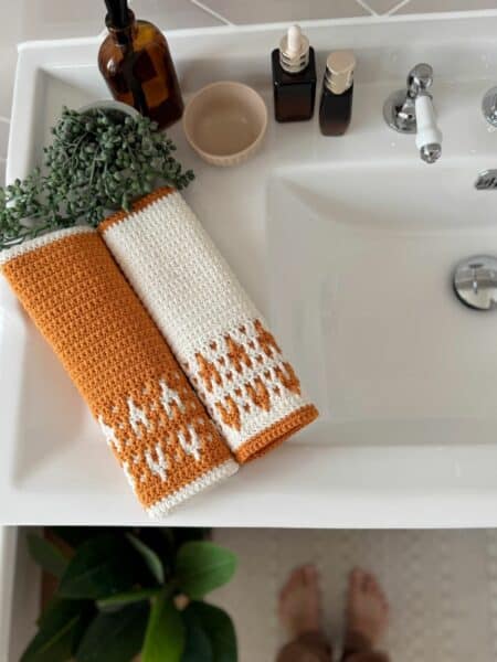 Two crocheted hand towels, one orange and one white with orange accents in an inset mosaic crochet design, are neatly placed on a white bathroom sink next to bottles and a small bowl.