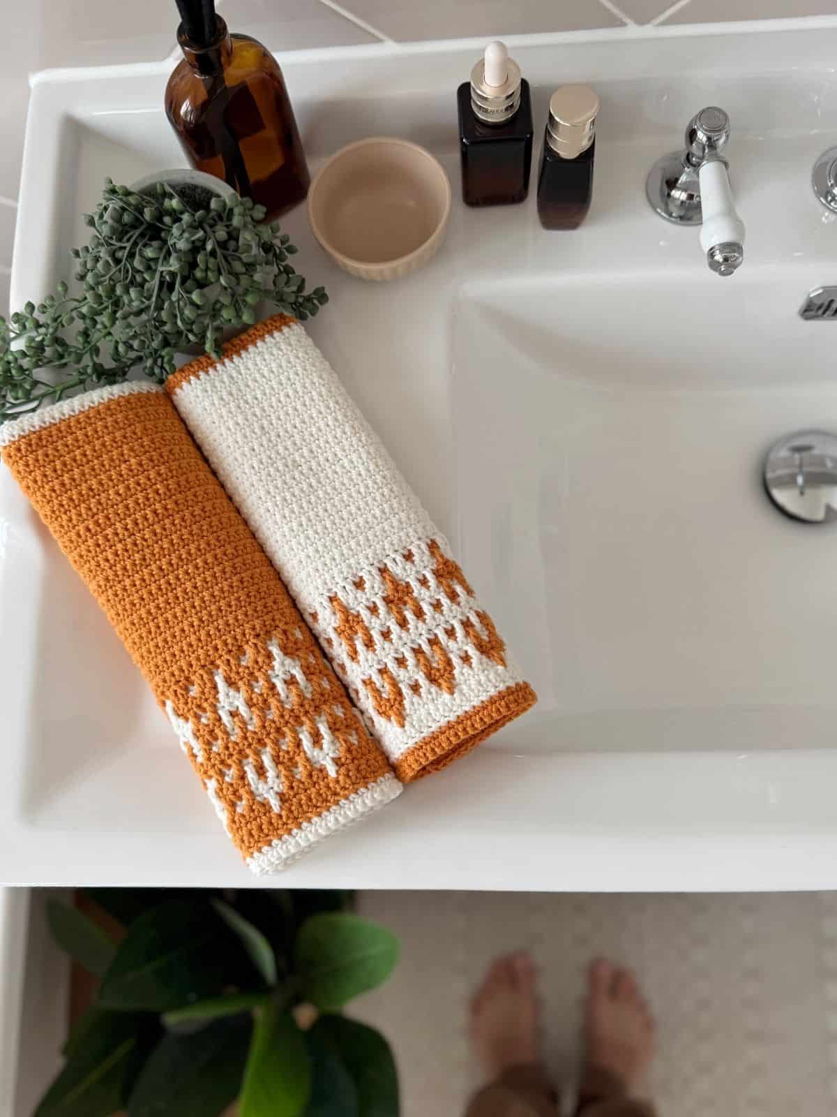 Two crocheted hand towels, one orange and one white with orange accents in an inset mosaic crochet design, are neatly placed on a white bathroom sink next to bottles and a small bowl.