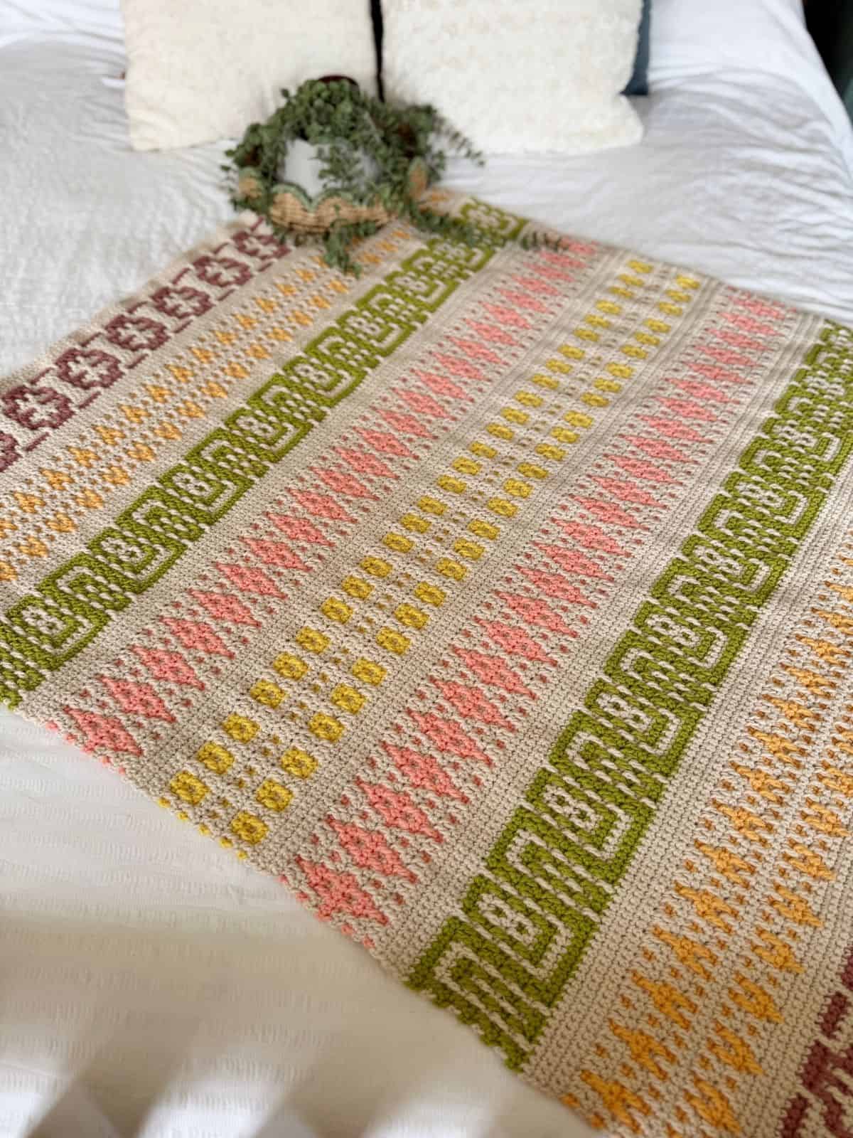 A colorful mosaic crochet blanket pattern with geometric designs in pink, green, yellow, and brown lies on a white bed with two pillows and a small plant.