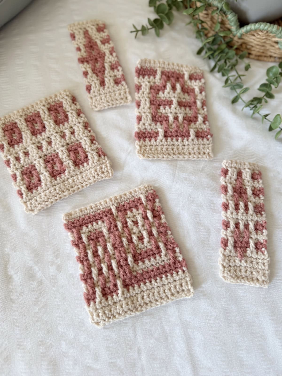Five rectangular crochet swatches with geometric patterns in cream and pink yarn are displayed on a white textured surface alongside eucalyptus leaves, perfect inspiration for your next mosaic crochet tutorial.