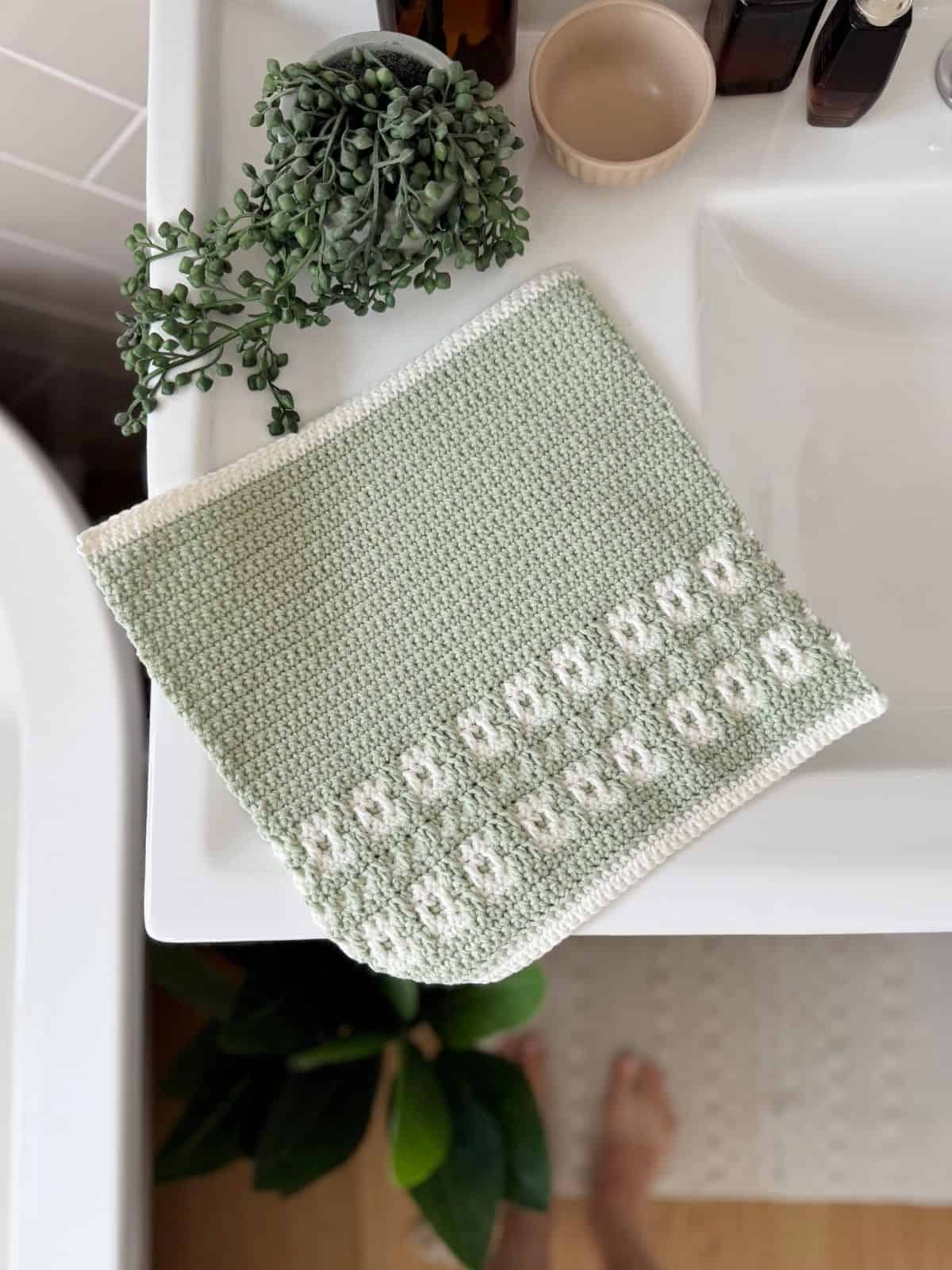 A crocheted green and white washcloth with a floral pattern, made from a mosaic crochet free pattern, lies on a bathroom sink next to a potted plant and toiletries.