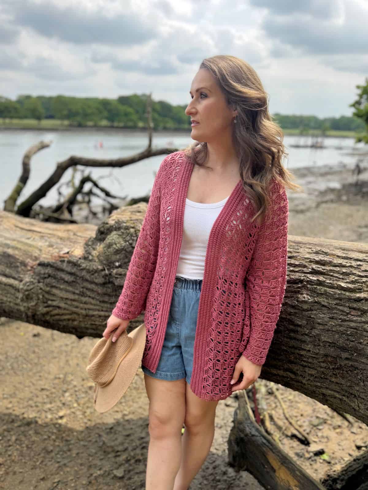 A woman in a pink summer cardigan crochet pattern and denim shorts stands by a large fallen tree near a riverside, holding a hat and looking to the side.