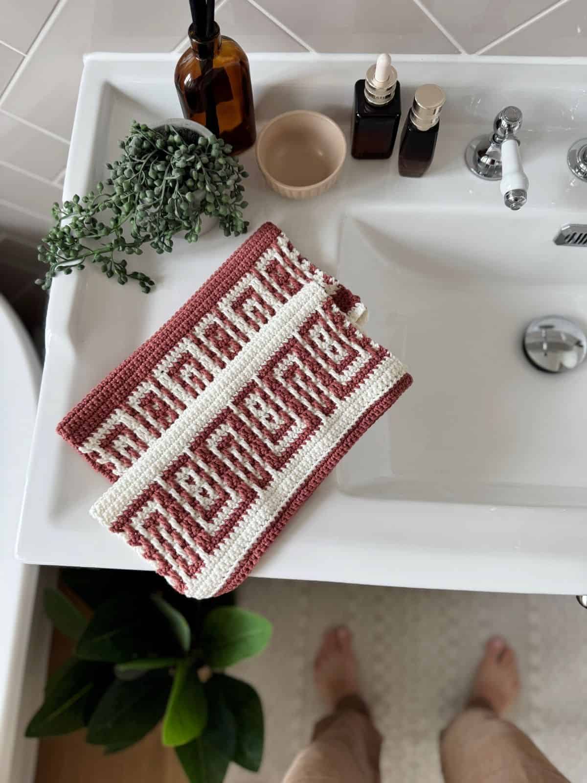 A bathroom sink with bottles, a plant, and a beige cup on the counter. A red and white patterned cloth, reminiscent of a free mosaic crochet pattern, is draped over the sink. A pair of feet is visible at the bottom of the image.